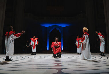 Five dancers in white robes with red stoles surrounding two people seated back to back in the middle of a labyrinth who are wearing red robes and have gold halos around their heads.