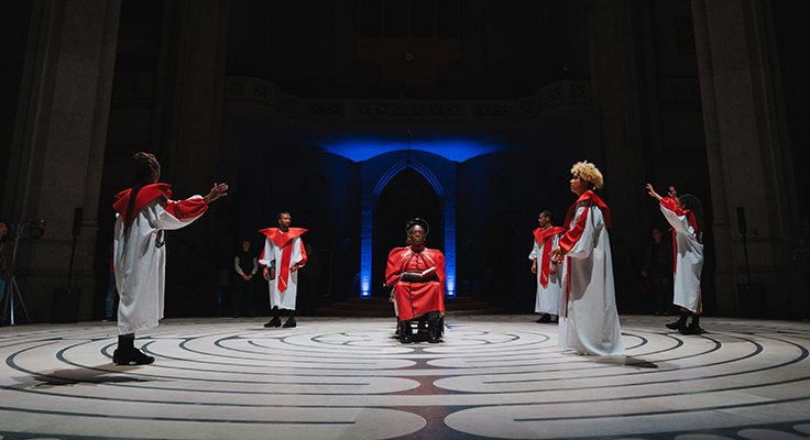 Five dancers in white robes with red stoles surrounding two people seated back to back in the middle of a labyrinth who are wearing red robes and have gold halos around their heads.