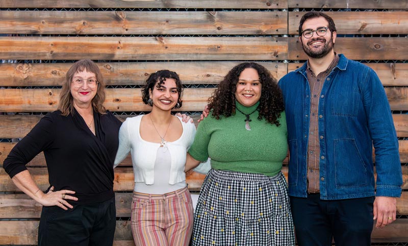 Four people stand close together, posing with their arms around each other’s backs.