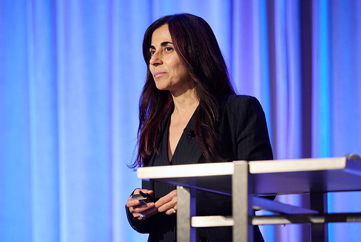 A person in a black blazer and with long black hair lightly grins and looks at and an audience that is not in the frame.