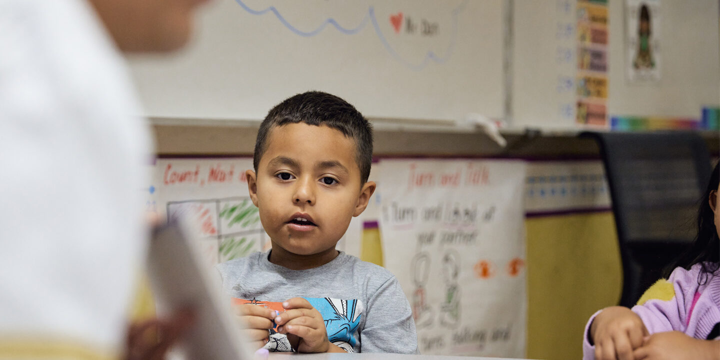 A young student looks intently at a flashcard that an out of focus teacher is holding.