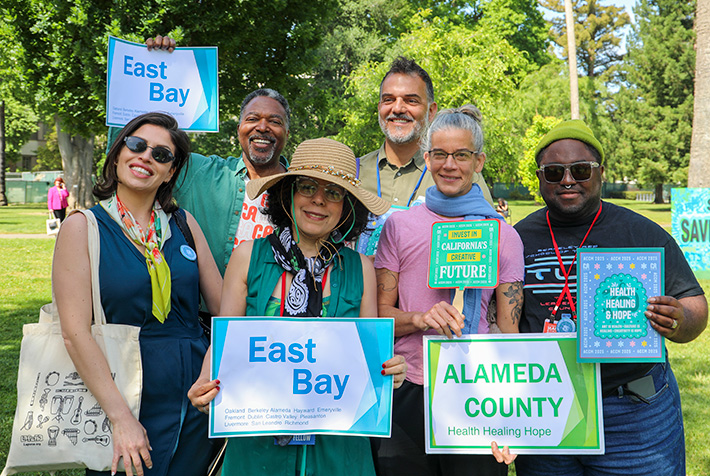 Six people stand closely together in a park. They are smiling and holding signs that say "East Bay," "Alameda County," "Invest in California's Creative Future," and "Health Healing and Hope."