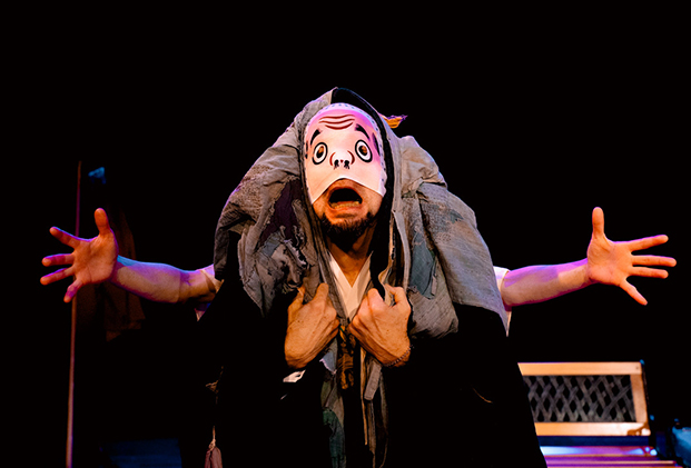 Two performers are entwined and look as one person with four arms—two extended outward and two clutching the chest. An expressive mask covers the nose and mouth and their mouth is wide open creating an animated facial expression.