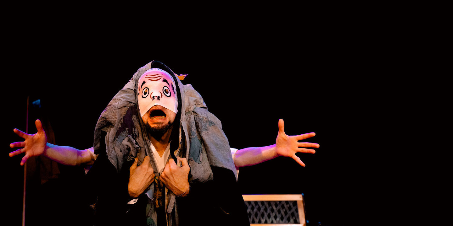 Two performers are entwined and look as one person with four arms—two extended outward and two clutching the chest. An expressive mask covers the nose and mouth and their mouth is wide open creating an animated facial expression.