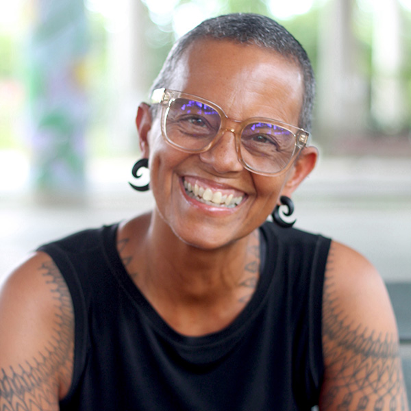 Sarah, a mixed race Black woman with very short salt and pepper hair, black spiral earrings, black tank top, and shoulder tattoos, smiles brightly at the camera. She is in front of a blurred background.