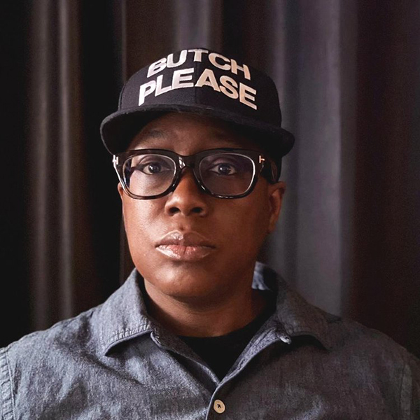 A Black person wearing glasses and a black baseball hat with white letters that read "Butch Please" looks directly at the camera.