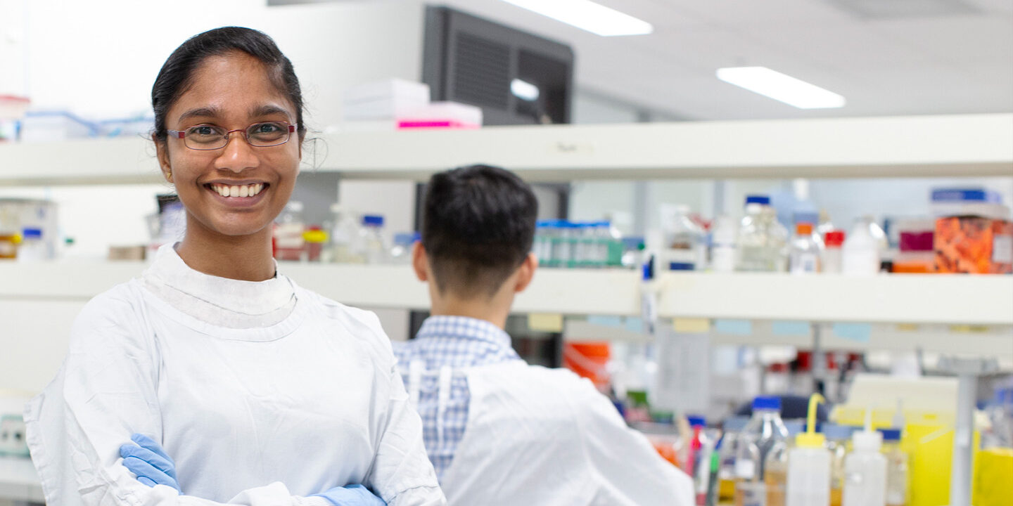 person smiles at camera in the background are shelves filled with medical lab supplies
