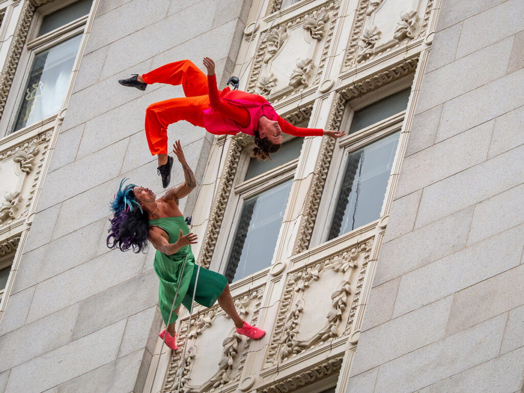 Two aerial dancers performing mid-air alongside a building with ornate windows.
