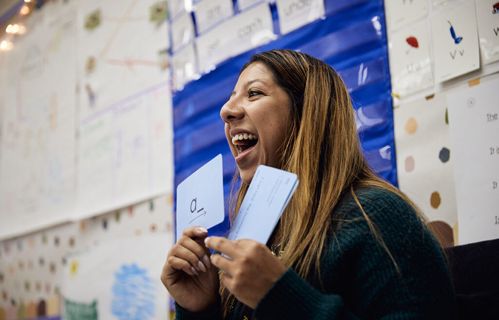 A teacher with a big smile on her face, holding up a card with the letter "a" and a blank space next to it for the students to fill in with other letters.
