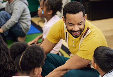 An educator sitting on the floor, in conversation with students.