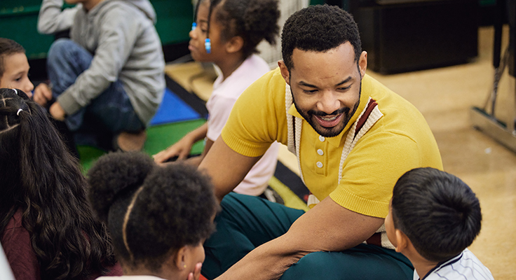 An educator sitting on the floor, in conversation with students.