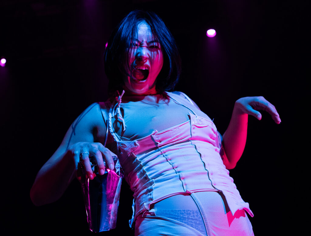 Performer wearing a nude colored tank top with vertical stitches, in one hand she holds a chrome colored cup while her other hand is draped down like a t-rex. She is lip syncing and her expression is challenging and defiant.