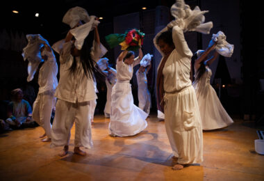 Eight dancers, all wearing white garments, are moving on stage. They're holding sheets of fabric above their heads.