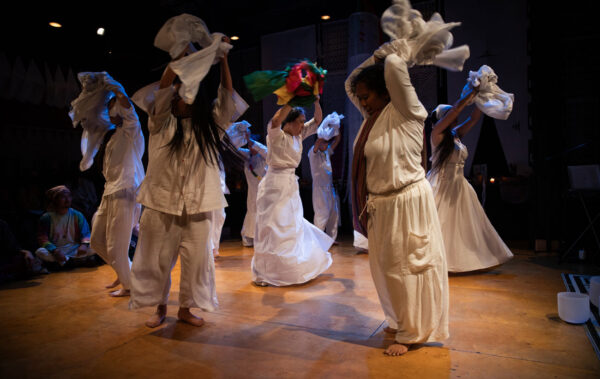 Eight dancers, all wearing white garments, are moving on stage. They're holding sheets of fabric above their heads.