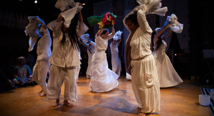 Eight dancers, all wearing white garments, are moving on stage. They're holding sheets of fabric above their heads.