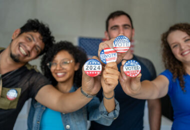 four smiling people hold up red, white and blue buttons that read "VOTE"
