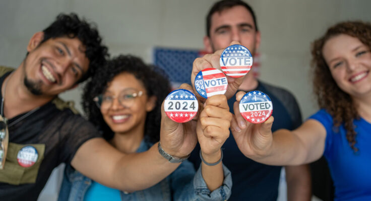 four smiling people hold up red, white and blue buttons that read "VOTE"