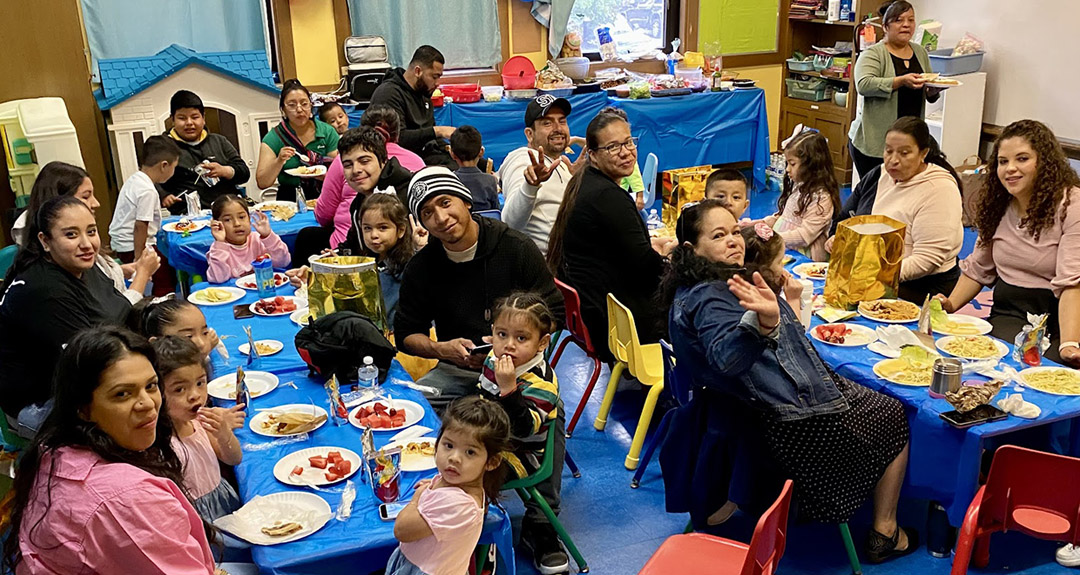 families and children are seated at long tables in child-size chairs. They are turned and smiling at the camera The tables are full of plates of food.