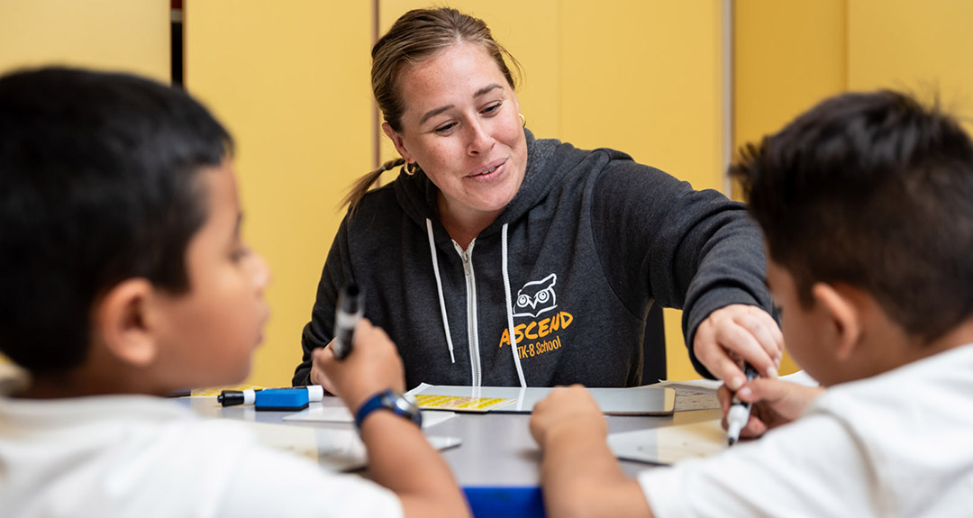 a teacher smiles while working with two elementary age students at a small table.