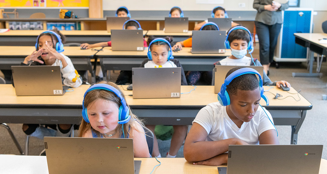 ten students wearing bright blue headphones look at laptops while seated at long tables in a classroom.