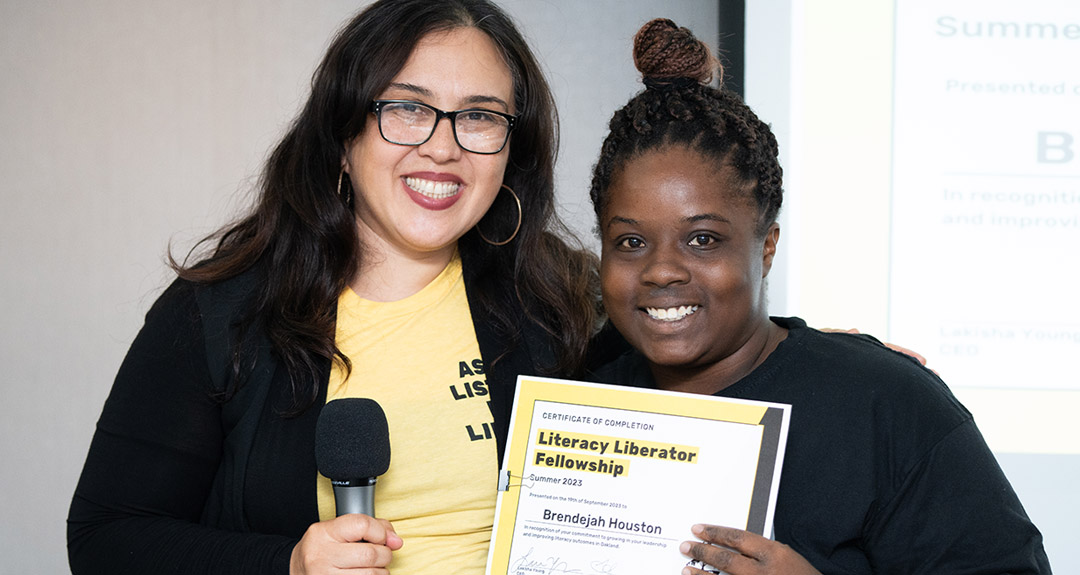 two people stand closely and smile at the camera. The person on the right is holding a Literacy Liberator Fellowship certificate