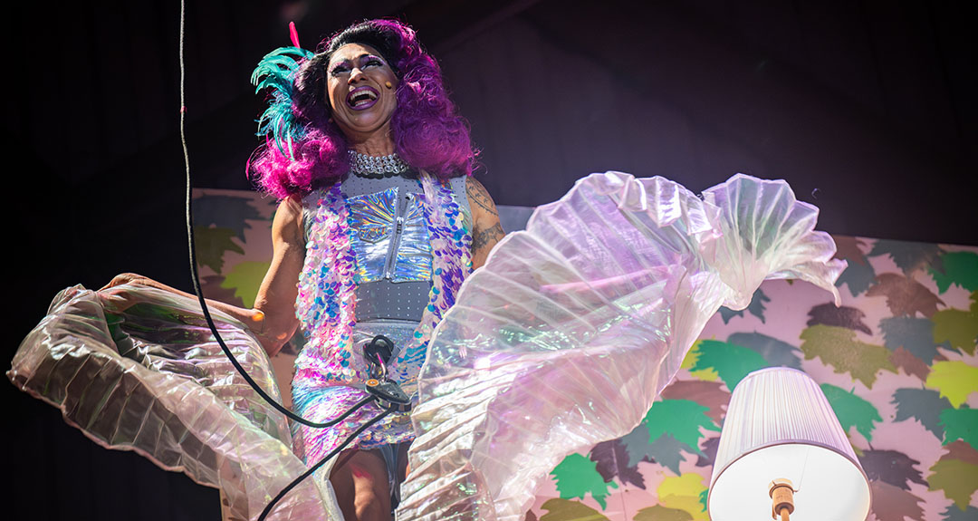 A person with purple hair and lipstick and wearing a multi-colored sequin dress holds a length of pleated iridescent fabric as it captures air. The person is attached to a rope and harness.