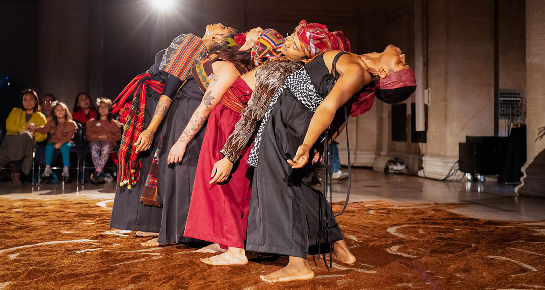 Four dancers bend backward against each other in a line. They stand barefoot on a stage covered in rust brown dirt.