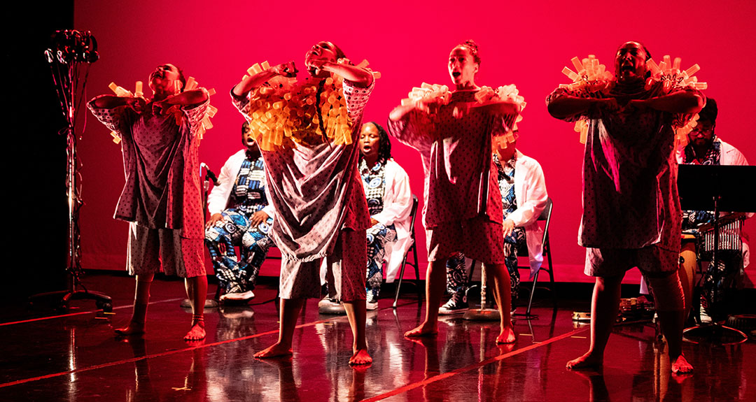 A stage bathed in red light. Four performers in white jackets and bold printed garments sing, while others are standing wearing uniquely crafted garments. Their shoulders are adorned with bright orange prescription bottles that to catch the light in a striking, artful display.