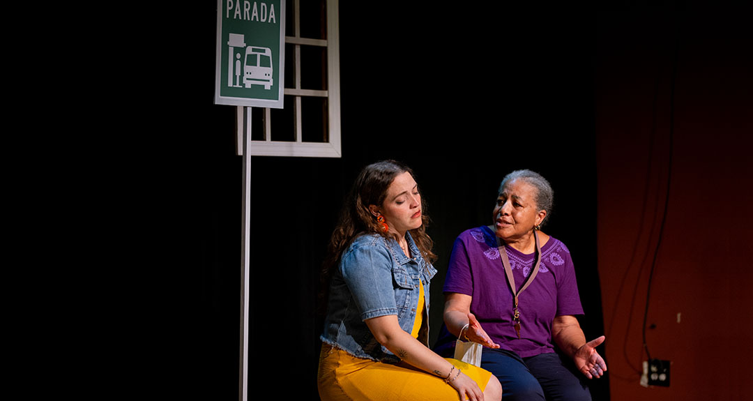 Two people sit next to each other on a bench and talk. A sign above them says "Parada" and has a bus icon.