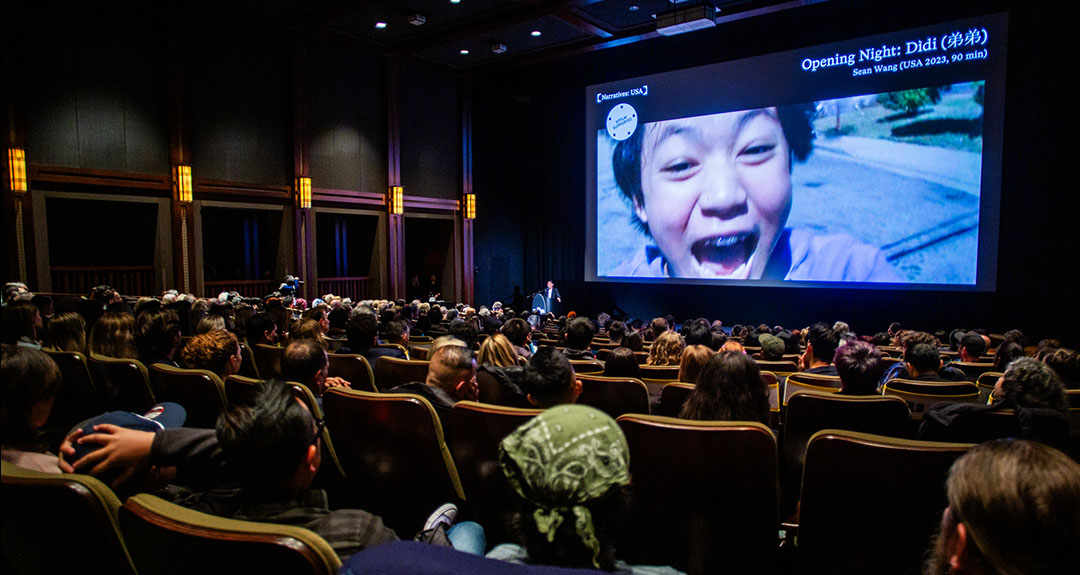 A full audience watches a film in a theater. On the screen a close-up of a young boy is projected.