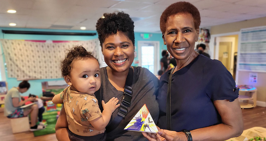 2024_TurningPoints_EDUC_AfroPlay Two adults smile at the camera; one holds a baby while the other holds a triangle-shaped object covered in colorful paint splotches. Behind them is a large room with adults and children playing together.