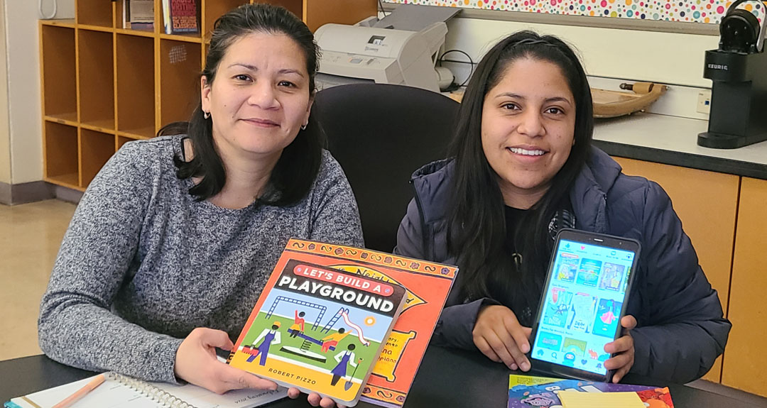 2024_TurningPoints_EDUC_AlwaysDream Two seated adults smile at the camera, each holding an item toward it. One displays a children's book titled Let's Build a Playground, while the other holds up a tablet showing a screen of tiny books.