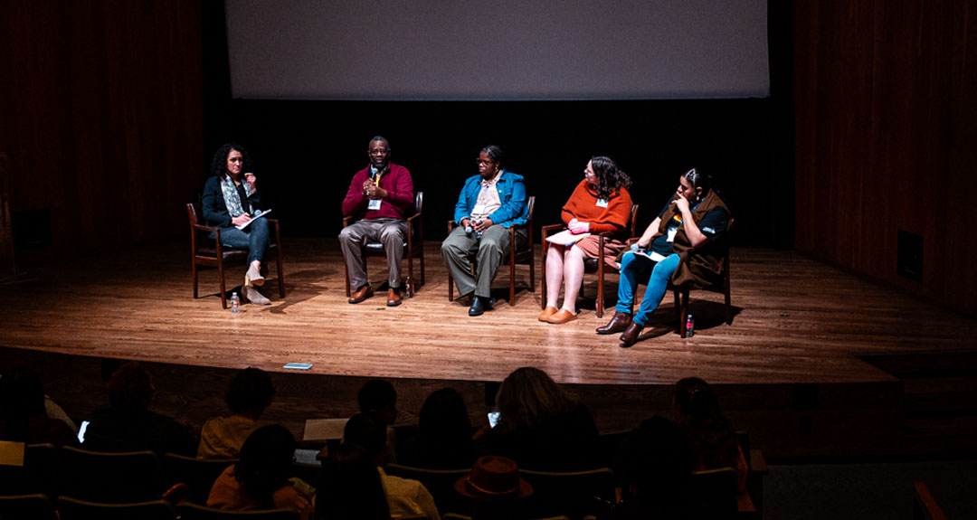 2024_TurningPoints_EDUC_RightToRead Five people are spotlighted and seated on a stage. One person is speaking into a microphone. In the background there is a white, blank projection screen. In the foreground an is audience in shadow.
