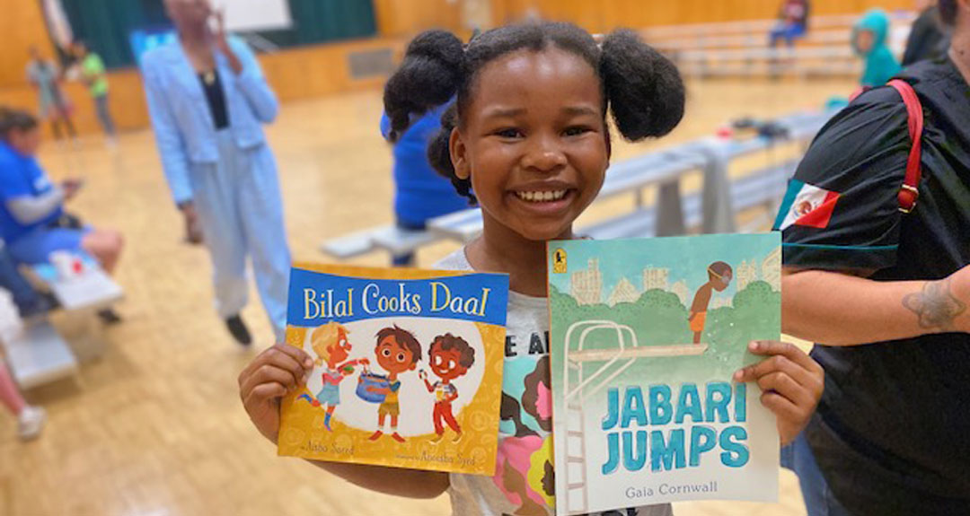 A young child with pig tails grins widely holding two books—"Bilal Cooks Daal" and "Jabari Jumps."