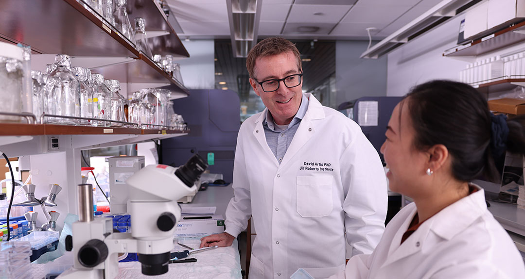 Two people smile at each other. They are wearing white coats and are standing in a scientific lab next to a microscope.