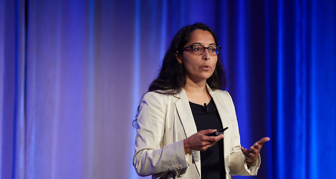 A person with long hair and wearing a white blazer and glasses speaks to an audience that is not visible in the image.