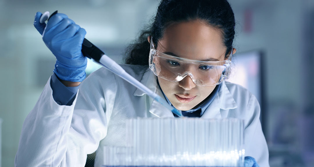 A person wearing safety goggles uses a pipette to fill test tubes.