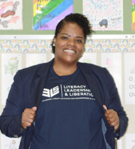 Smiling person stands against a classroom wall of drawings. They are wearing a dark blue blazer and matching t-shirt printed with white logo reading Literacy, Leadership and Liberation